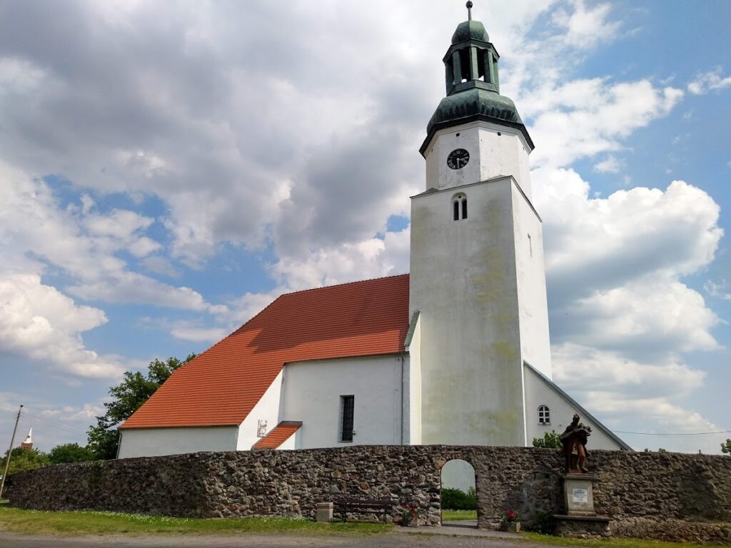 Biały budynek z wieżą kościelną, pomarańczowym dachem. Wokół mur z kamienia i figura.