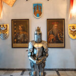 Jedna z sal w zamku. Na pierwszym planie trzy zbroje rycerskie. za nimi na ścianie wiszą obrazy przodków.