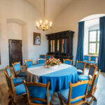 Odrestaurowany salon w zamku w Kożuchowie. Na srodku okrągły stół zasłany błękitnym obrusem, wokół krzesła obite błękitnym suknem. Po praekj stronie pokoju duże okno z błękitnymi zasłonami do ziemi, obok drewaniana, masywna komoda, dalej drewniane drzwi. Podłoga w kaflach.